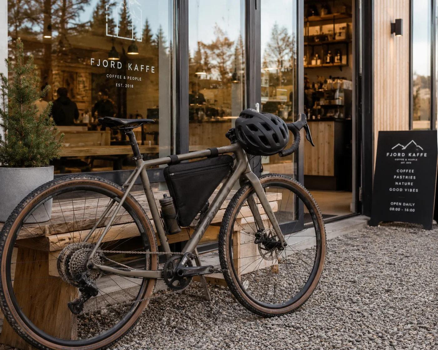Sand-coloured gravel bike parked on gravel outside a wood-and-glass coffee shop, a black frame bag strapped inside the frame triangle above the bottle and a helmet hung on the bars